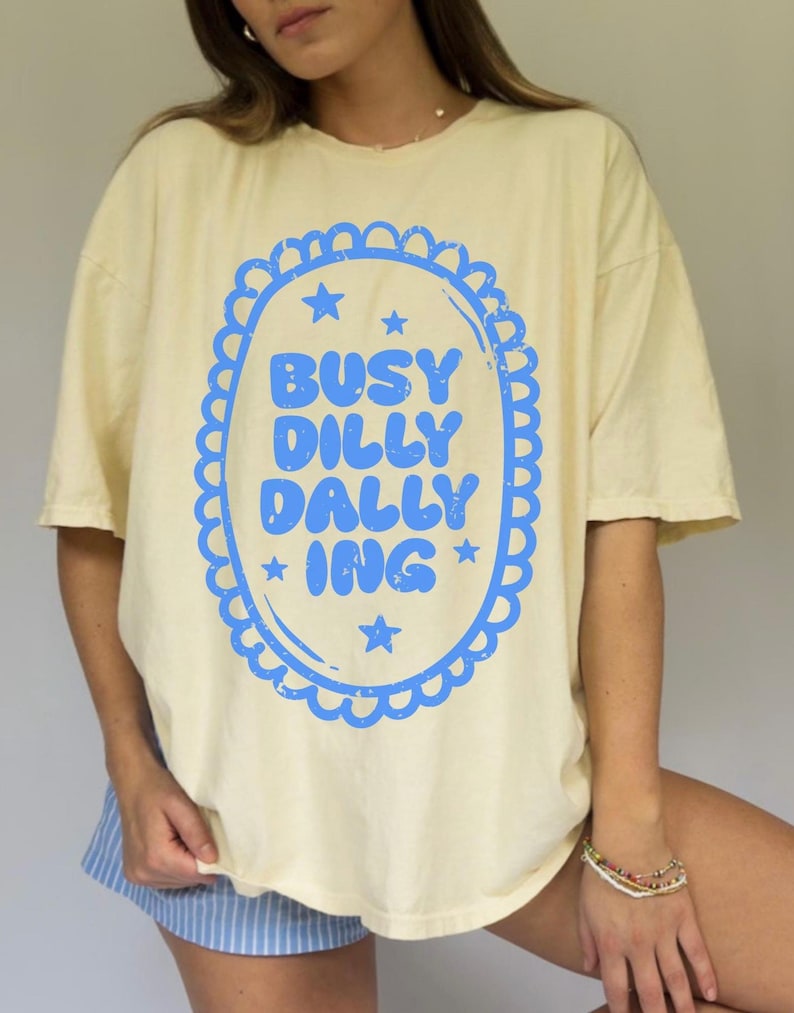May include: A pale yellow t-shirt featuring a blue oval design with the words "BUSY DILLY DALLY ING". The design is framed by a scalloped border and stars. The shirt has short sleeves.