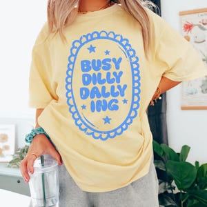 May include: A pale yellow t-shirt featuring the blue text "BUSY DILLY DALLY ING" within a blue scalloped oval with stars. The shirt has a relaxed fit.