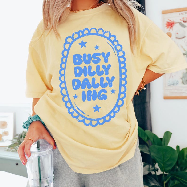 May include: A pale yellow t-shirt featuring the blue text "BUSY DILLY DALLY ING" within a blue scalloped oval with stars. The shirt has a relaxed fit.