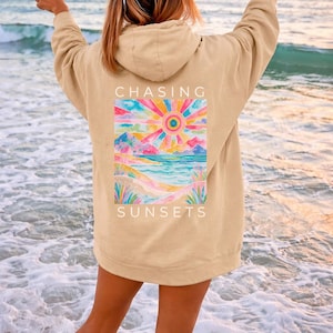 Op de afbeelding: Beige hoodie met een kleurrijke zonsondergangafbeelding op de rug. De afbeelding toont een levendige zon, bergen en een oceaansc&egrave;ne met de woorden "CHASING SUNSETS". De persoon staat op een strand met de armen in een vredesteken.