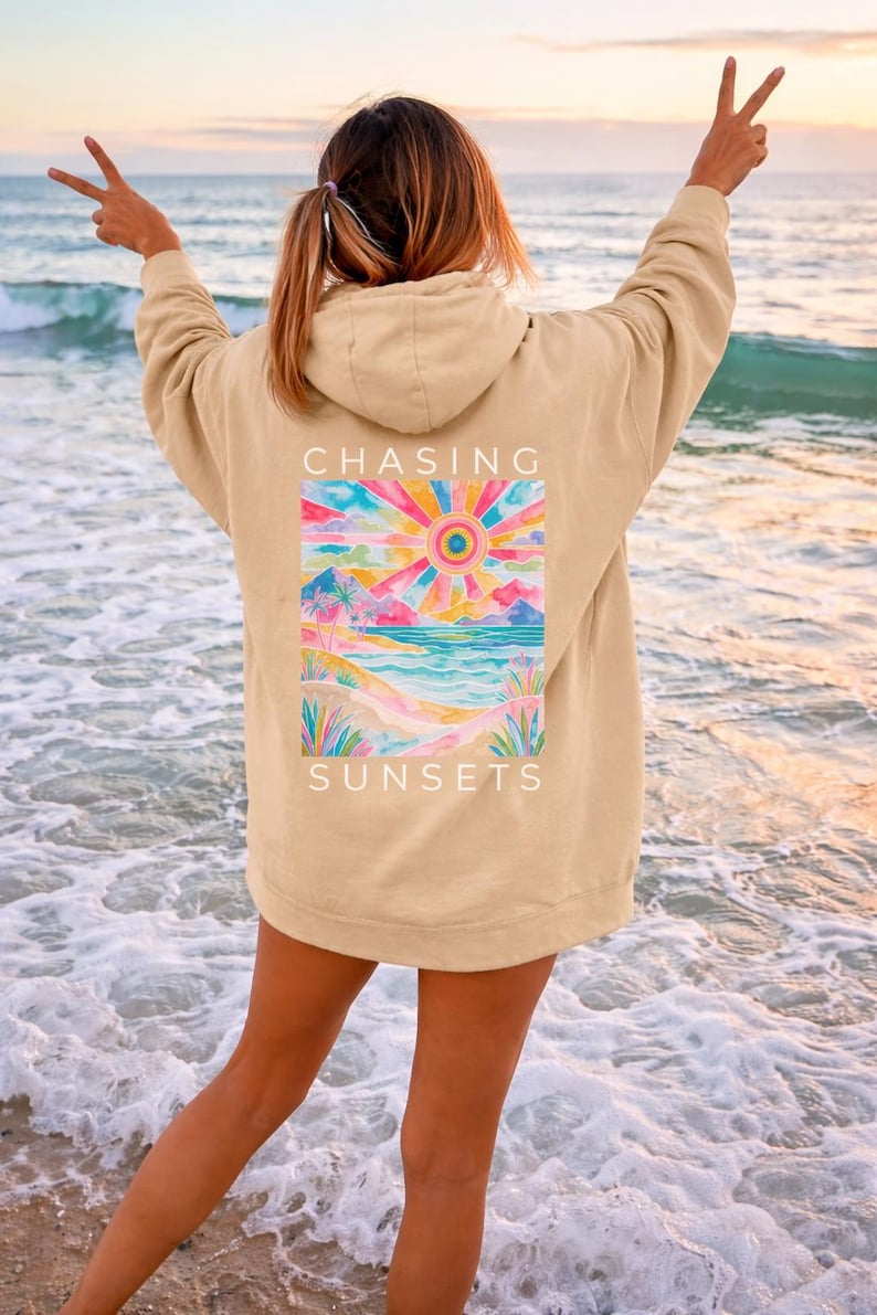 Op de afbeelding: Beige hoodie met een kleurrijke zonsondergangafbeelding op de rug. De afbeelding toont een levendige zon, bergen en een oceaansc&egrave;ne met de woorden "CHASING SUNSETS". De persoon staat op een strand met de armen in een vredesteken.