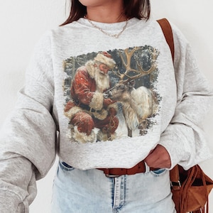 May include: A light gray crewneck sweatshirt featuring a graphic print of Santa Claus in a red suit with white fur trim, kneeling and interacting with a reindeer in a snowy forest setting. The sweatshirt has long sleeves and a ribbed neckline.