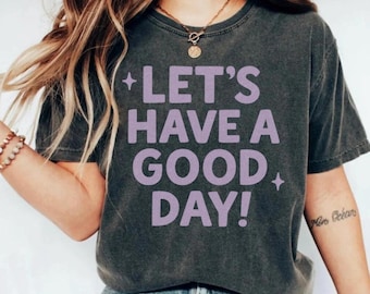 Może przedstawiać: Ciemnoszara koszulka z napisem "LET'S HAVE A GOOD DAY!" w kolorze lawendowym. Koszulka ma luźny krój i jest wykonana z miękkiego materiału. Tekst otoczony jest gwiazdkami.