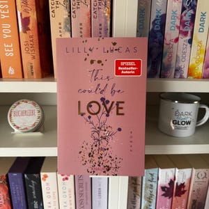 Könnte beinhalten: Ein rosa Buch mit dem Titel "This Could Be Love" von Lilly Lucas, mit goldfarbenen Buchstaben und floralen Akzenten. Das Buch wird auf einem weißen Regal, umgeben von anderen Büchern, ausgestellt. Eine Tasse mit "Dark Glow" ist sichtbar.