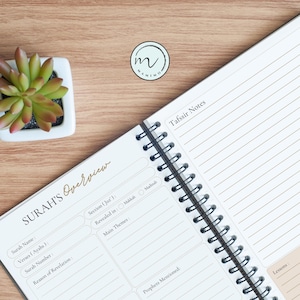 May include: A spiral-bound notebook with white pages featuring printed sections for Surah's Overview and Tafsir Notes. The layout includes fields for Surah Name, Verses, Section, and Main Themes. A small succulent in a white square pot is nearby.