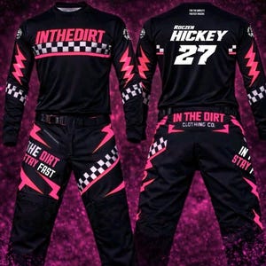 Könnte beinhalten: Ein schwarzes Rennset aus Langarmtrikot und Hose mit pinkfarbenen Blitzmotiven und weißen Karomustern. Das Trikot zeigt den Text INTHEDIRT, ROCZEN HICKEY 27 und 2026 RACE KIT. Die Hose ist mit dem Schriftzug THE DIRT STAY FAST versehen.
