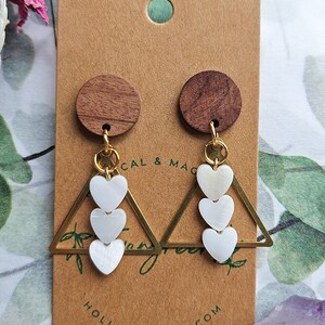 May include: A pair of gold triangle earrings with three white heart-shaped charms hanging from each. The earrings are displayed on a brown card with the text "HOLLYAXELROD.COM" printed on it.