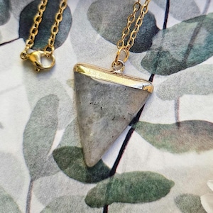 Puede incluir: Un collar de cadena dorada con un colgante triangular de piedra gris. El colgante tiene un borde dorado.