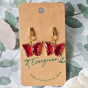 May include: A pair of red butterfly charm earrings with gold accents and clear rhinestones. The earrings are on a brown card with the text "Evergreen" and "hollyaxelrod.com".