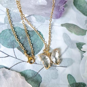 Puede incluir: Un collar de cadena dorada con un colgante de mariposa. El colgante de mariposa está hecho de cristales transparentes y facetados. El collar se muestra sobre una superficie estampada con cristales.