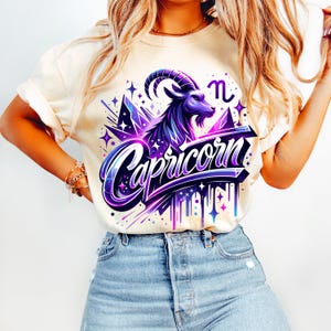 Peut inclure: Un t-shirt blanc avec un motif graphique violet et bleu repr&eacute;sentant un signe du zodiaque Capricorne avec le mot "Capricorne" dans une police stylis&eacute;e. Le motif comprend &eacute;galement des &eacute;toiles et un effet de goutte &agrave; goutte.