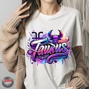 Peut inclure: T-shirt blanc avec un motif graphique color&eacute; repr&eacute;sentant le mot "Taurus" dans une police stylis&eacute;e avec une t&ecirc;te de taureau violette et un symbole astrologique.