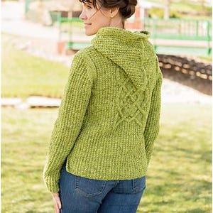 Könnte beinhalten: Ein grüner Kapuzenpullover mit strukturiertem Strickmuster und einem dekorativen Muster auf dem Rücken. Der Pullover wird mit einer blauen Jeans kombiniert. Das Bild wurde im Freien bei natürlichem Licht aufgenommen.