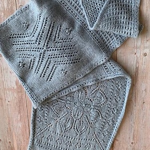 May include: A light gray knitted scarf with intricate openwork and floral patterns. The scarf features a detailed design with a geometric starburst pattern on one end and a floral motif on the other. The scarf is made of soft yarn.