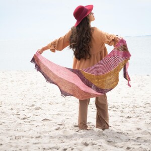 May include: A colourful shawl with a striped and gradient design. The shawl features shades of pink, gold, and purple. The shawl has a scalloped edge. The person is wearing a burgundy hat and a tan dress.