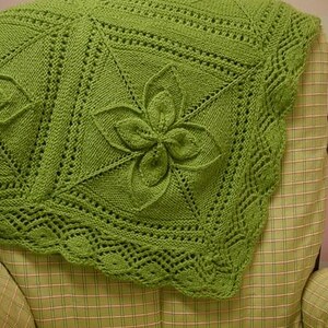 May include: A handmade, green knitted blanket with a floral design. The blanket features raised flower motifs and intricate openwork patterns. The edges have a scalloped border. The blanket is draped over a plaid armchair.