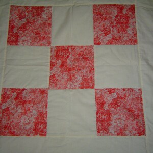May include: A handmade quilt featuring a checkerboard pattern. The quilt has alternating squares of cream-colored fabric and squares with a red floral print. The quilt is likely made of cotton and has a simple, classic design.