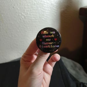 May include: A circular black pin-back button featuring a pixelated rainbow-colored text that reads Ask me about my Hyper-fixations. The design includes small red heart icons and white star accents scattered across the dark background surface.