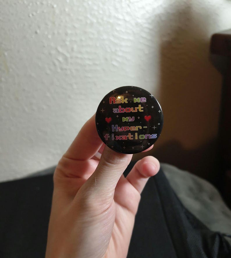 May include: A circular black pin-back button featuring a pixelated rainbow-colored text that reads Ask me about my Hyper-fixations. The design includes small red heart icons and white star accents scattered across the dark background surface.
