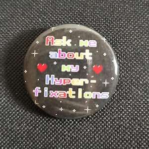Puede incluir: Una chapa redonda con fondo negro que presenta destellos blancos y dos corazones rojos. El texto Ask me about MY Hyper-fixations aparece en una fuente pixelada multicolor. El diseño está centrado en la superficie de la chapa circular.