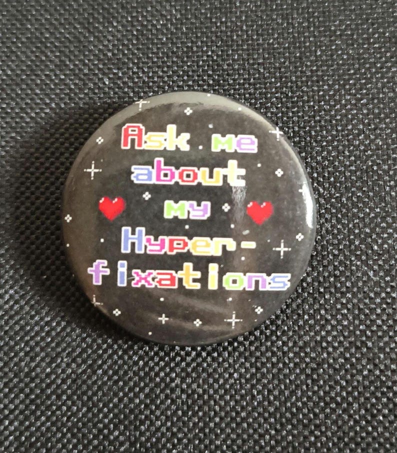 May include: A circular pin-back button with a black background featuring white sparkles and two red hearts. The text reads Ask me about MY Hyper-fixations in a multi-colored, pixelated font style. The design is centered on the round button surface.