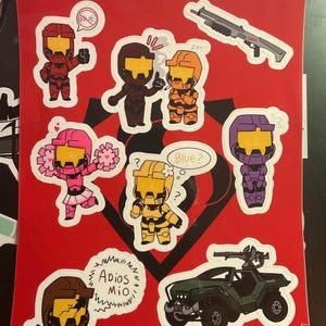 May include: A collection of cartoon stickers on a red background, featuring characters in various colors, including red, brown, orange, pink, yellow, and purple. The stickers depict figures in armor, weapons, and vehicles. Text includes "RED TEAM" and speech bubbles.