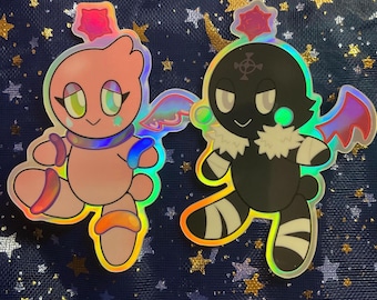 Op de afbeelding: Twee holografische stickers met stripfiguren. Eén is roze met een stervormige hoofdtooi, vleugels en een sjaal. De andere is zwart met een witte kraag, vleugels en een kruissymbool. Beide hebben een schattig ontwerp.