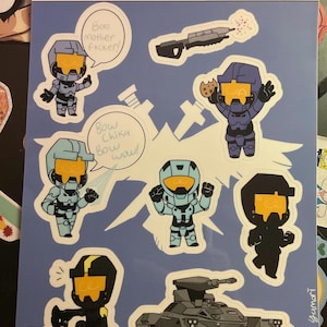 May include: A sheet of stickers featuring cartoon characters in futuristic armor with orange visors. The stickers are in various colors, including blue and black, and some have speech bubbles with text. The words "BLUE TEAM" are written at the top.