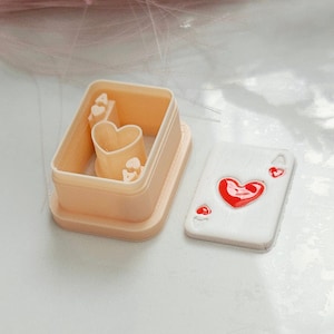 May include: A light peach-colored cookie cutter set with a heart theme. The set includes a rectangular frame with heart-shaped cutouts and a separate card-shaped piece with a red heart design. Ideal for baking themed cookies.