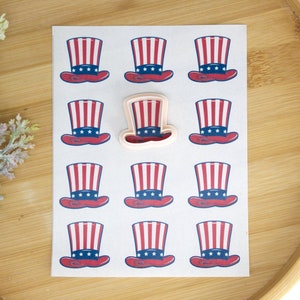 May include: A sheet of white paper with 12 red, white, and blue Uncle Sam hat cookie cutters. One of the cookie cutters is a plastic template.