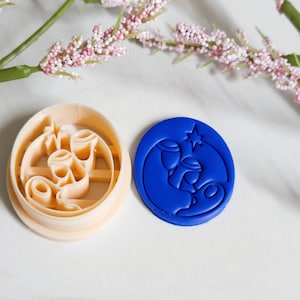 May include: Two cookie cutters. One is beige and oval-shaped with a design of three candles. The other is a blue, circular cutter with a nativity scene design. The cutters are on a white surface with pink flowers in the background.