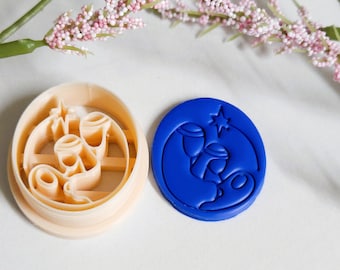 May include: Two cookie cutters. One is beige and oval-shaped with a design of three candles. The other is a blue, circular cutter with a nativity scene design. The cutters are on a white surface with pink flowers in the background.