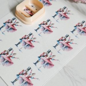 May include: A sheet of paper with repeating watercolor illustrations of a ballerina and a nutcracker soldier. The ballerina wears a pink tutu and the soldier has a blue coat. A beige cookie cutter is in the upper left corner.