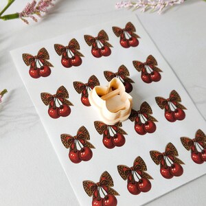 May include: A sheet of cherry stickers with leopard print bows and a beige cookie cutter in the shape of a cat. The cherries are red with brown stems and the bows are brown and tan.