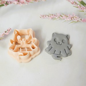 May include: A light peach-colored cat-shaped cookie cutter next to a gray cat-shaped cookie. The cookie cutter has a detailed design, while the cookie has a simple, cute face. The items are on a white surface with pink floral accents.