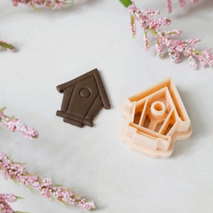 May include: A set of two brown and pink plastic cookie cutters shaped like birdhouses. The cutters are on a white surface.