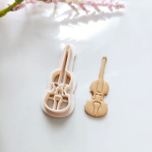 May include: A white plastic cookie cutter and a brown cookie in the shape of a violin.