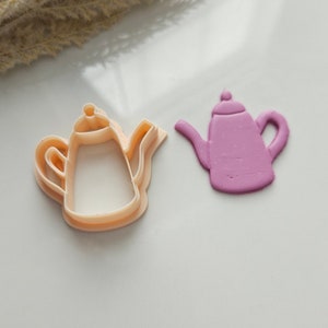 May include: Two teapot cookie cutters, one is pink and the other is beige. The beige cutter is a cutout design and the pink cutter is solid.