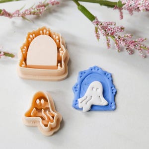 May include: Three Halloween-themed cookie cutters. One is a ghost shape, another is a ghost inside a blue frame, and the third is a church-like shape. The cutters are in shades of beige and blue.