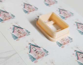 May include: A light peach-colored rectangular cookie cutter sits on a sheet of paper with repeating watercolor illustrations of pink houses and blue trees. The cutter has a rectangular shape with a hollow center.