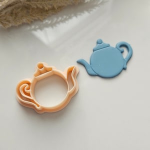 May include: Two teapot cookie cutters, one is light blue and the other is a light peach color. The cookie cutters are on a white surface.