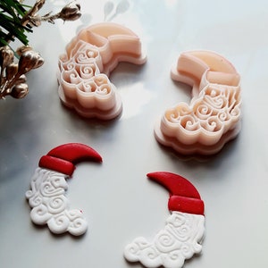 May include: Four Santa Claus-shaped cookie cutters and two Santa Claus-shaped fondant decorations. The cutters are light pink, and the decorations are white with red hats. A sprig of greenery and gold-colored buds are in the background.
