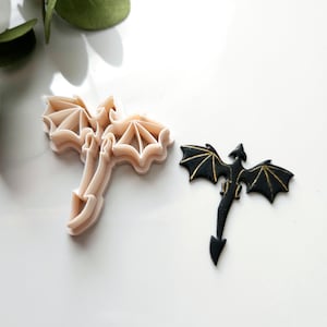 May include: Two dragon cookie cutters, one white and one black. The white cutter is a detailed silhouette of a dragon with wings spread. The black cutter is a solid dragon with gold accents.