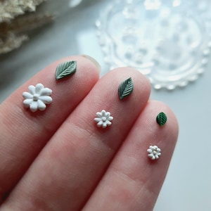 May include: A hand holding five small polymer clay charms: two white flowers, two green leaves, and one green leaf with a white flower.