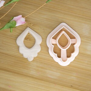 May include: Two earring-shaped polymer clay cutouts and a matching cutter sit on a wooden surface. The clay cutout on the left is a light beige color, while the cutter on the right is a light pink. A pink flower and green stem are in the upper left corner.