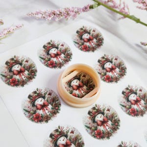 May include: A sheet of round stickers featuring a watercolor snowman design. The snowman wears a top hat and a red bow, surrounded by pinecones and red flowers. A small, beige container holds a matching sticker.