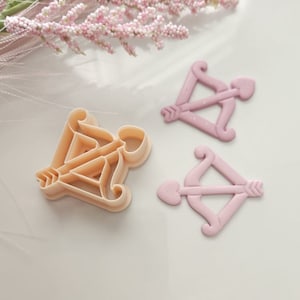 May include: Two pink and beige plastic cookie cutters shaped like an arrow and bow. The bow is shaped like a heart.