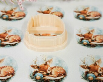 May include: A beige plastic cookie cutter with a rectangular opening, surrounded by several printed images of a sleeping deer with antlers, a blue ornament, and pine cones. The deer images are on a white background.