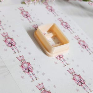 May include: A light peach-colored rectangular cookie cutter with interior dividers sits on a patterned paper. The paper features repeating illustrations of pink nutcracker soldiers and snowflakes, creating a festive holiday theme.