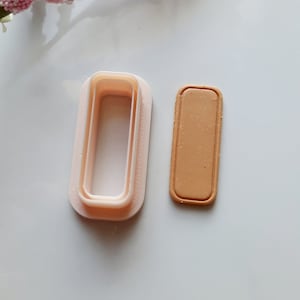 May include: A set of two plastic cookie cutters, one is a rectangle with a smaller rectangle inside, the other is a solid rectangle. Both are light pink in color.